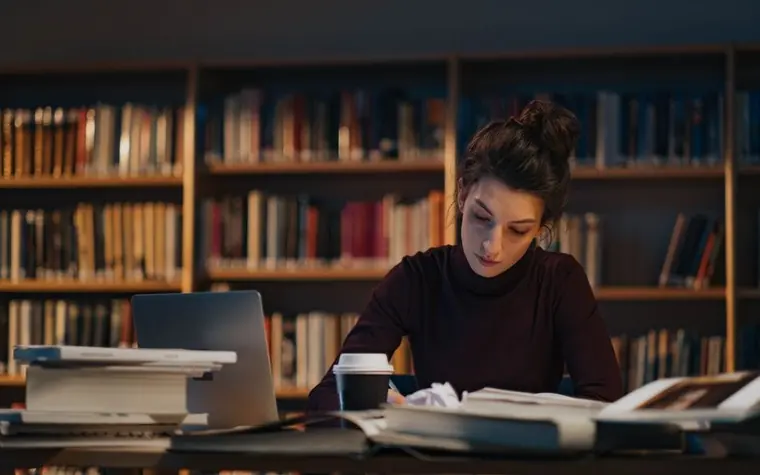 An exam candidate studying at a library desk with a stack of books