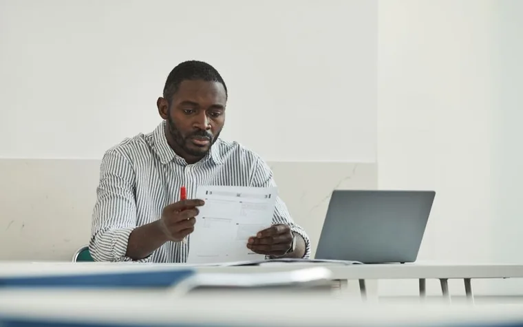 A man at a laptop looking over a test paper with a red pen in hand