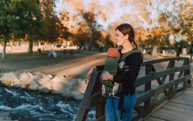 A mother carrying a baby in a carrier while standing on a wooden bridge in a park