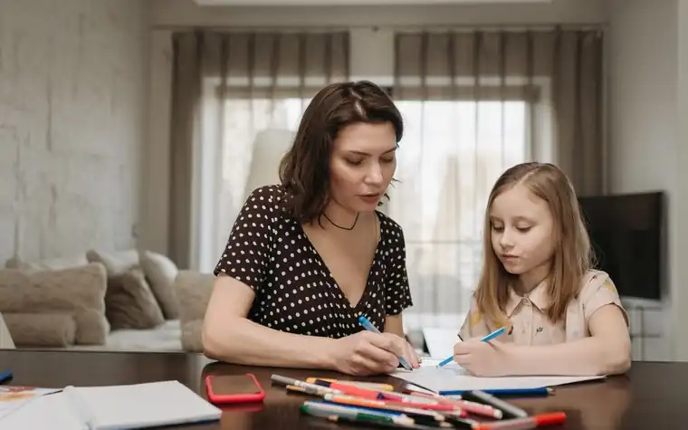 A child drawing with colored pencils at a table, with a guardian writing beside them