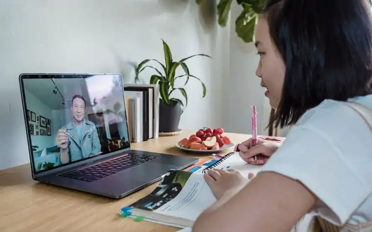 A girl taking notes in a notebook while watching a laptop video class