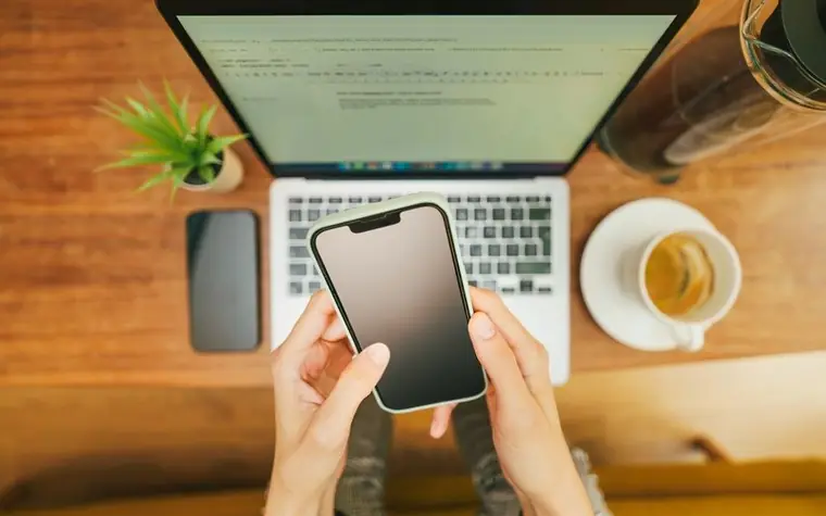 A hand holding a smartphone in front of a laptop on a cafe table while checking a document