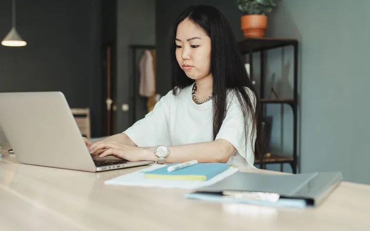 A woman working on a laptop at a bright desk with documents and books beside her