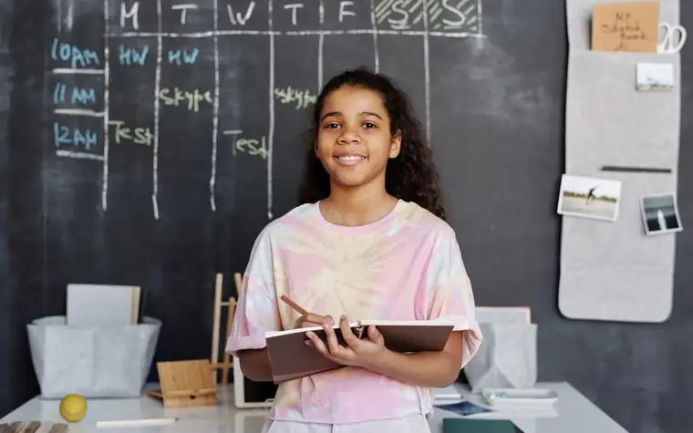 A student smiling with a notebook in front of a timetable on a chalkboard
