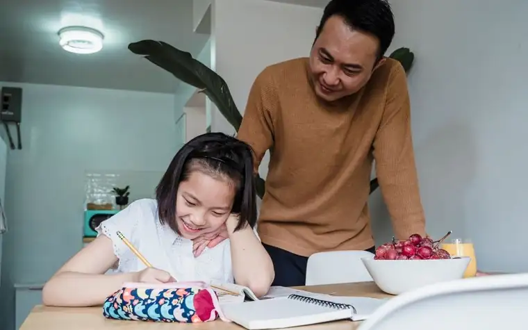 A father watching his daughter do homework at the dining table