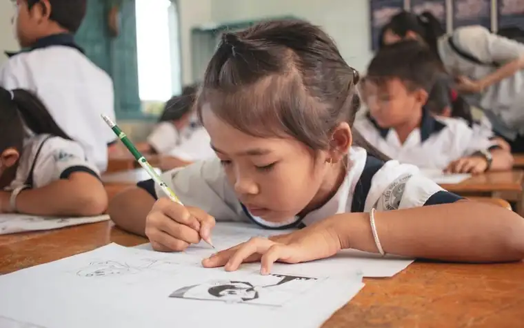 A schoolchild bending over a desk drawing with a pencil, with classmates in the classroom